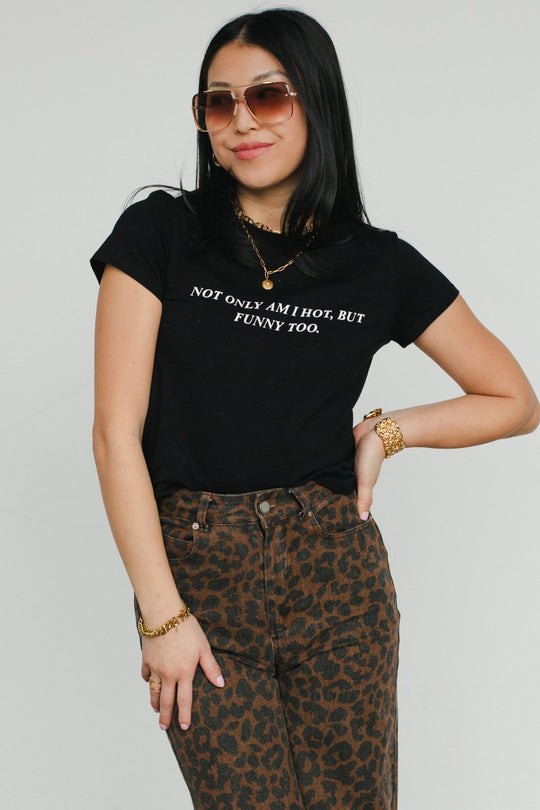 A woman poses confidently against a plain white background, wearing Panache Apparel Collection’s Not Only Am I Hot, But Funny Too Baby Tee with sunglasses and high-waisted leopard print pants.