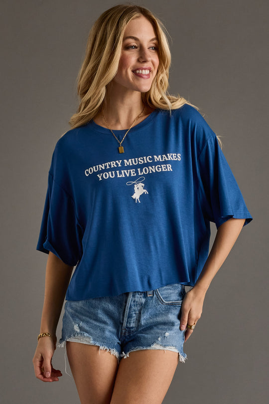 A smiling woman with long blonde hair wears the Panache Apparel Collection's Country Music Cropped Tee and distressed denim shorts, standing against a plain gray background.