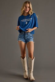 A woman stands against a gray background, smiling and holding her Panache Apparel Collection Country Music Cropped Tee with the slogan “Country Music Makes You Live Longer,” paired with denim shorts and white cowboy boots.