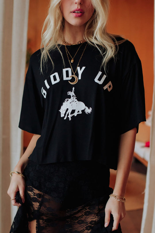 A woman stands indoors in soft lighting, wearing the Panache Apparel Collection's Giddy Up Cropped Tee with a cowboy graphic, layered necklaces, and a sheer black lace skirt.