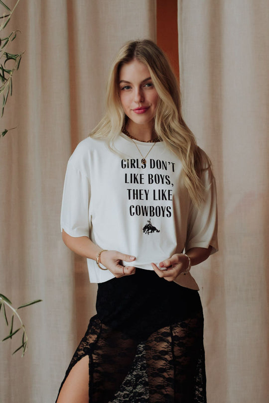 A woman with long blonde hair stands indoors, smiling as she holds the hem of her Girls Don't Like Boys Cropped Tee by Panache Apparel Collection—styled with a high-slit black lace skirt for a modern western look.
