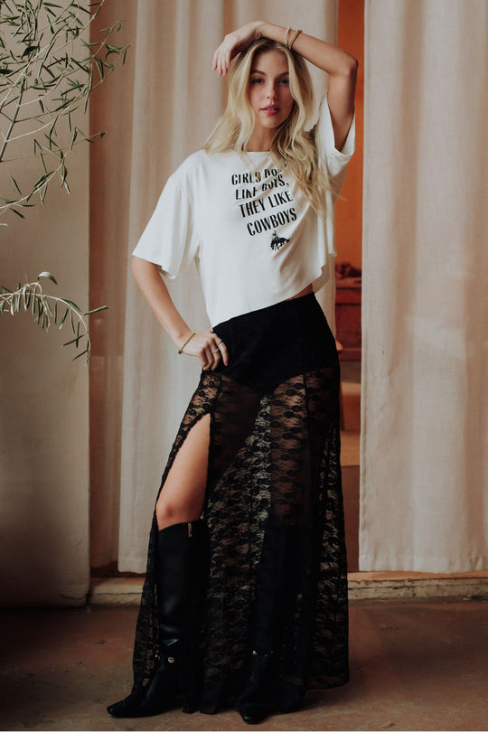 A woman with long blonde hair poses indoors in the Girls Don't Like Boys Cropped Tee by Panache Apparel Collection, tucked into a high-waisted black lace skirt with thigh-high slits and paired with black knee-high boots in front of cream curtains.