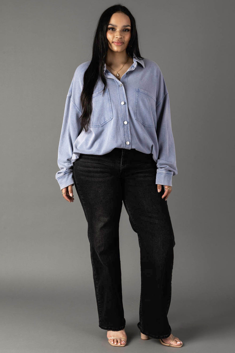 A woman with long dark hair and a confident expression wears the Panache Apparel Collection Light Blue Washed Soft Button Up—an oversized fit top with large front pockets—paired with black jeans and clear open-toed heels against a gray background.