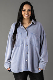 A woman with long dark hair smiles in the Panache Apparel Collection Light Blue Washed Soft Button Up, featuring an oversized fit and two large chest pockets, paired with black pants against a plain gray background.