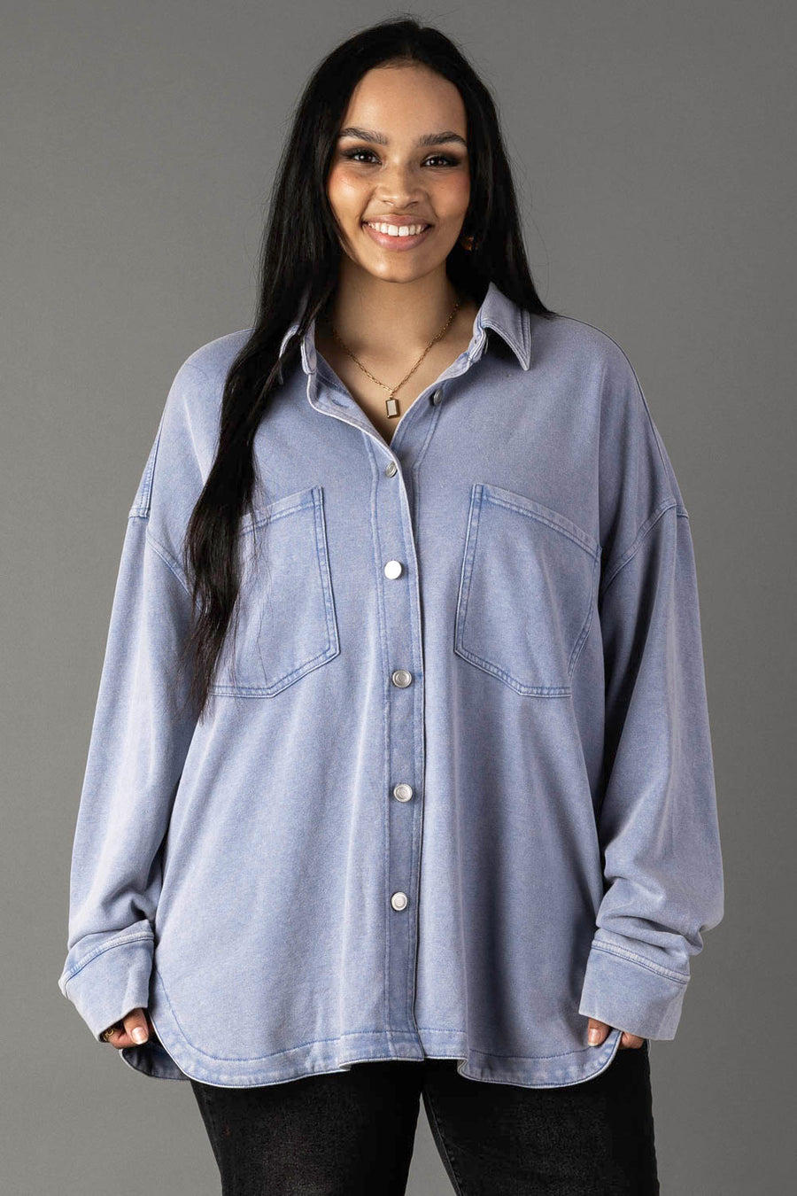 A woman with long dark hair smiles in the Panache Apparel Collection Light Blue Washed Soft Button Up, featuring an oversized fit and two large chest pockets, paired with black pants against a plain gray background.
