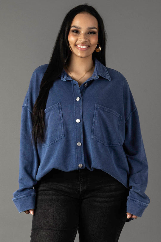 A woman with long dark hair wears the Panache Apparel Collection's Dark Blue Washed Soft Button Up, styled oversized with two front pockets and black jeans, standing and smiling against a plain gray background.