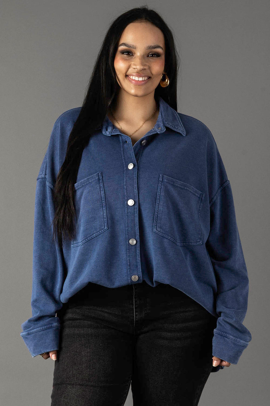 A woman with long dark hair wears the Panache Apparel Collection's Dark Blue Washed Soft Button Up, styled oversized with two front pockets and black jeans, standing and smiling against a plain gray background.