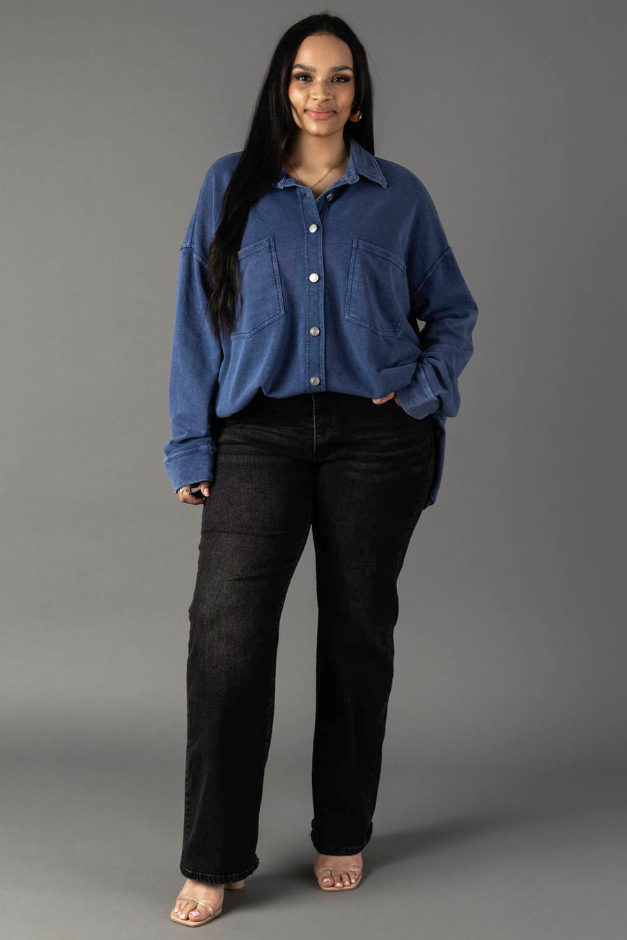 A woman with long dark hair stands against a gray background, wearing the Panache Apparel Collection Dark Blue Washed Soft Button Up, paired with black jeans and clear-heeled sandals. She smiles gently with one hand in her pocket.