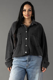 A woman with long dark hair models the Panache Apparel Collection Black Washed Soft Button Up—an oversized, cotton-blend shirt with large front pockets—paired with light blue jeans, standing against a plain gray background and smiling.