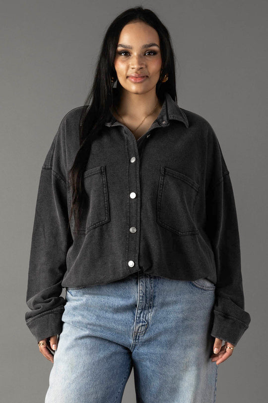 A woman with long dark hair models the Panache Apparel Collection Black Washed Soft Button Up—an oversized, cotton-blend shirt with large front pockets—paired with light blue jeans, standing against a plain gray background and smiling.