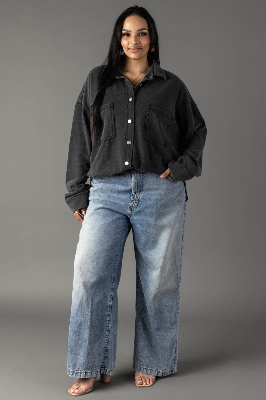 A woman with long dark hair smiles gently against a gray background, wearing the Panache Apparel Collection Black Washed Soft Button Up in an oversized fit and wide-leg light blue jeans, with one hand in her pocket.