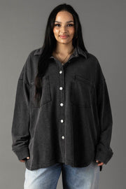 A woman with long dark hair wears the Panache Apparel Collection Black Washed Soft Button Up, featuring two chest pockets, paired with light blue jeans. She stands against a plain grey background and smiles softly at the camera.