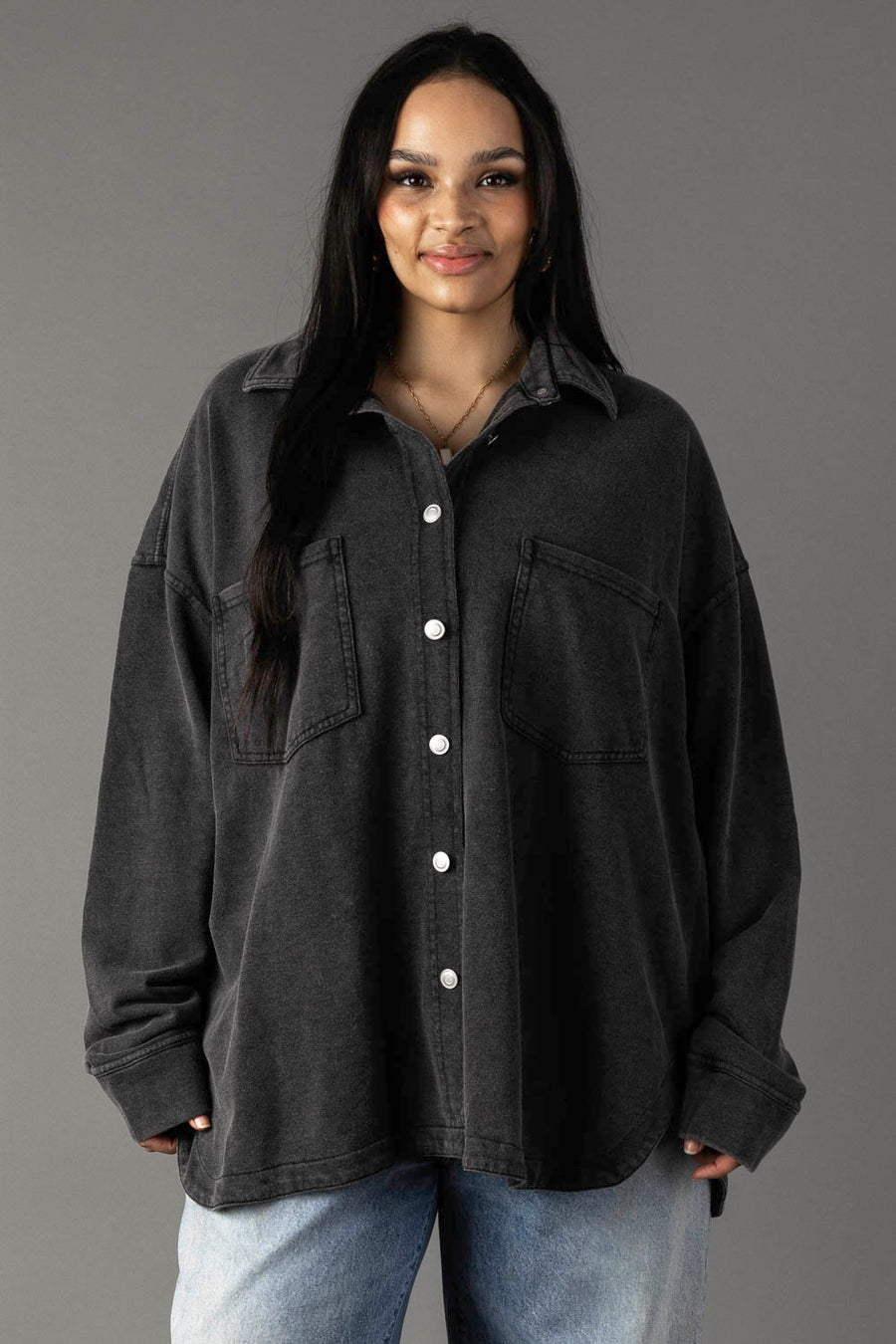 A woman with long dark hair wears the Panache Apparel Collection Black Washed Soft Button Up, featuring two chest pockets, paired with light blue jeans. She stands against a plain grey background and smiles softly at the camera.