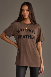 A woman with long dark hair wears the Panache Apparel Collection Whiskey Weather Tee in dark brown, styled with black pants and a gold necklace, standing against a plain gray background.