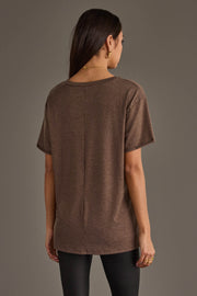 A woman with long dark hair, seen from behind, wears the Panache Apparel Collection Whiskey Weather Tee in dark brown and black pants against a plain gray background. She accessorizes with gold rings and a delicate necklace.
