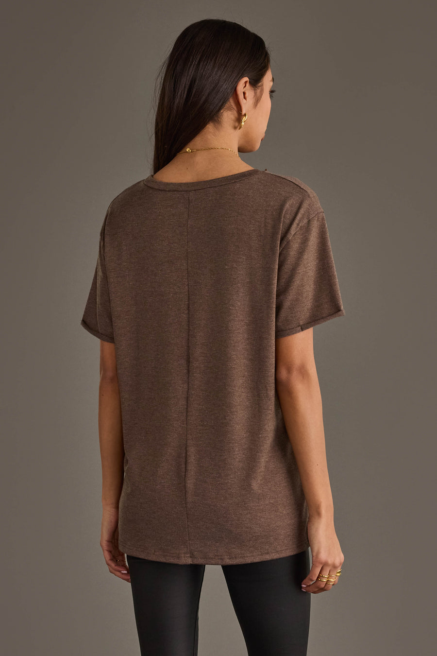 A woman with long dark hair, seen from behind, wears the Panache Apparel Collection Whiskey Weather Tee in dark brown and black pants against a plain gray background. She accessorizes with gold rings and a delicate necklace.