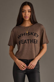 A woman with long dark hair models the Panache Apparel Collection Whiskey Weather Tee in dark brown, styled with black pants and gold jewelry, posing against a plain gray background.