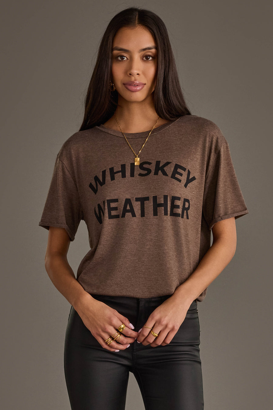 A woman with long dark hair models the Panache Apparel Collection Whiskey Weather Tee in dark brown, styled with black pants and gold jewelry, posing against a plain gray background.
