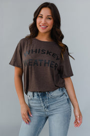 Woman wearing a brown t-shirt with 'WHISKEY WEATHER' text and light blue jeans against a gray background
