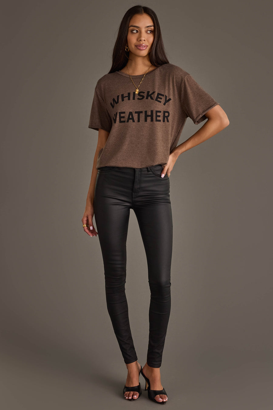 A woman poses against a plain background wearing the Panache Apparel Collection Whiskey Weather Tee in dark brown, paired with black skinny pants and heeled sandals. She has long hair and stands with one hand on her hip.
