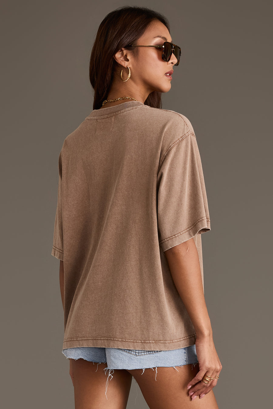 A woman wearing sunglasses and gold hoop earrings stands facing away, looking over her shoulder against a plain background. She is dressed in frayed denim shorts and the relaxed fit “Beer Weather Tee” by Panache Apparel Collection.