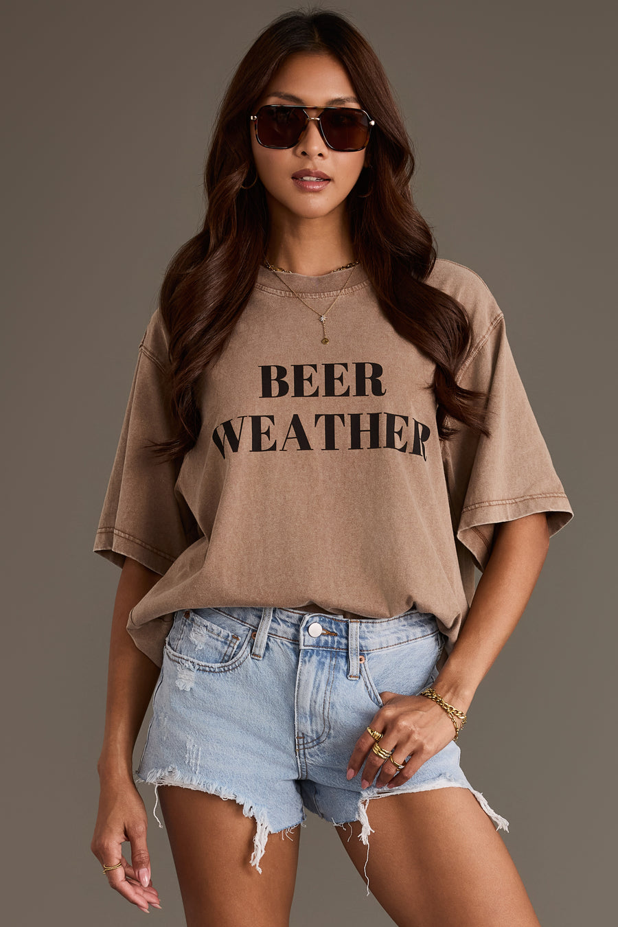 A woman with long wavy hair and sunglasses poses against a plain backdrop, wearing the Panache Apparel Collection’s Beer Weather Tee in relaxed fit brown cotton, paired with distressed denim shorts and gold jewelry.