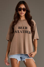A woman in the Panache Apparel Collection Beer Weather Tee—a relaxed-fit brown cotton t-shirt with “BEER WEATHER” printed on the front—styled with gold jewelry and denim shorts, standing against a plain background.