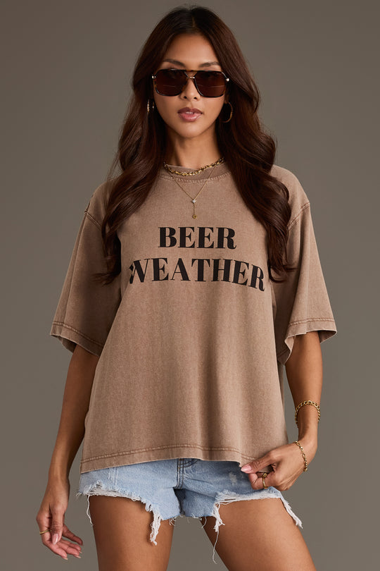 A woman in the Panache Apparel Collection Beer Weather Tee—a relaxed-fit brown cotton t-shirt with “BEER WEATHER” printed on the front—styled with gold jewelry and denim shorts, standing against a plain background.