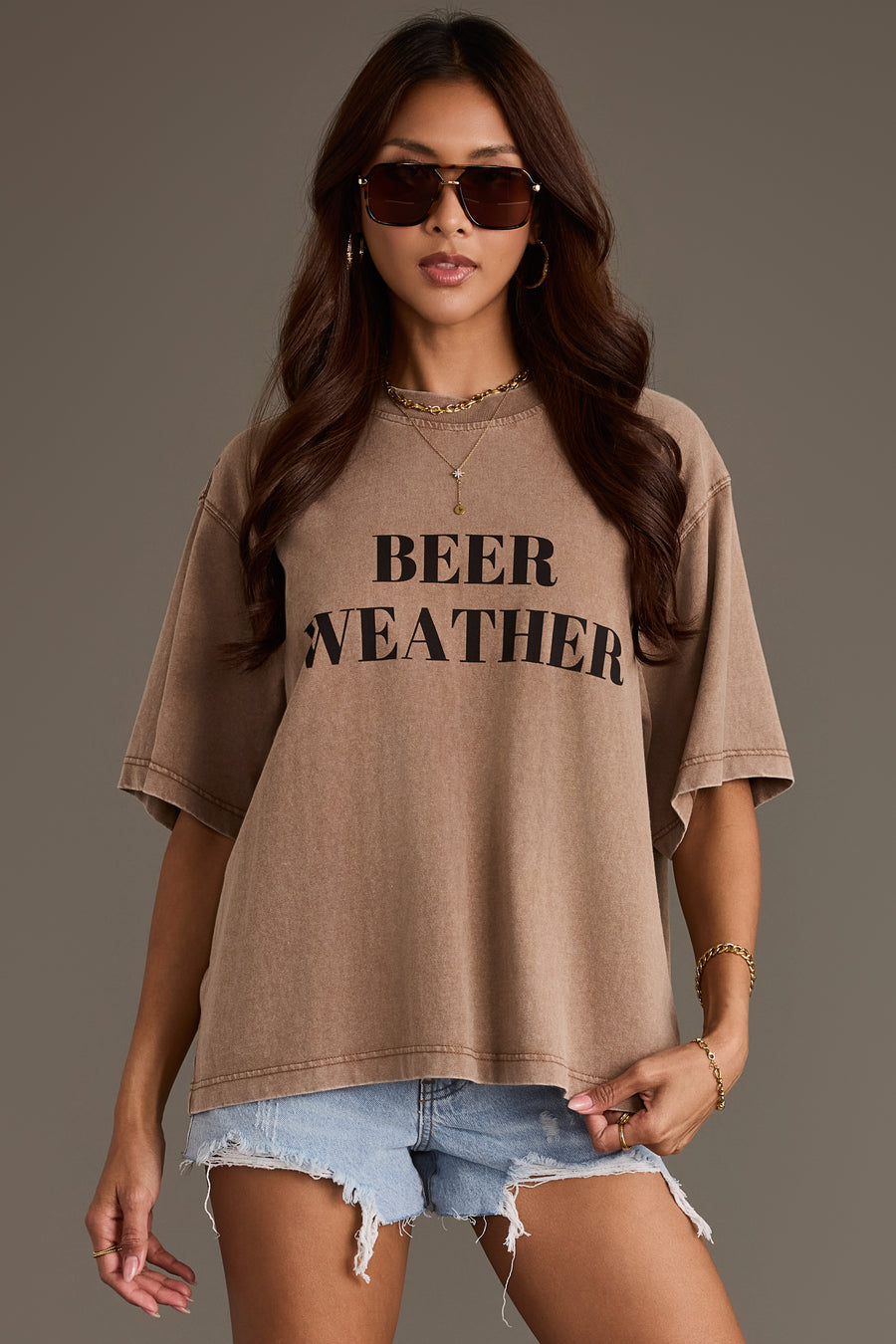 A woman in the Panache Apparel Collection Beer Weather Tee—a relaxed-fit brown cotton t-shirt with “BEER WEATHER” printed on the front—styled with gold jewelry and denim shorts, standing against a plain background.