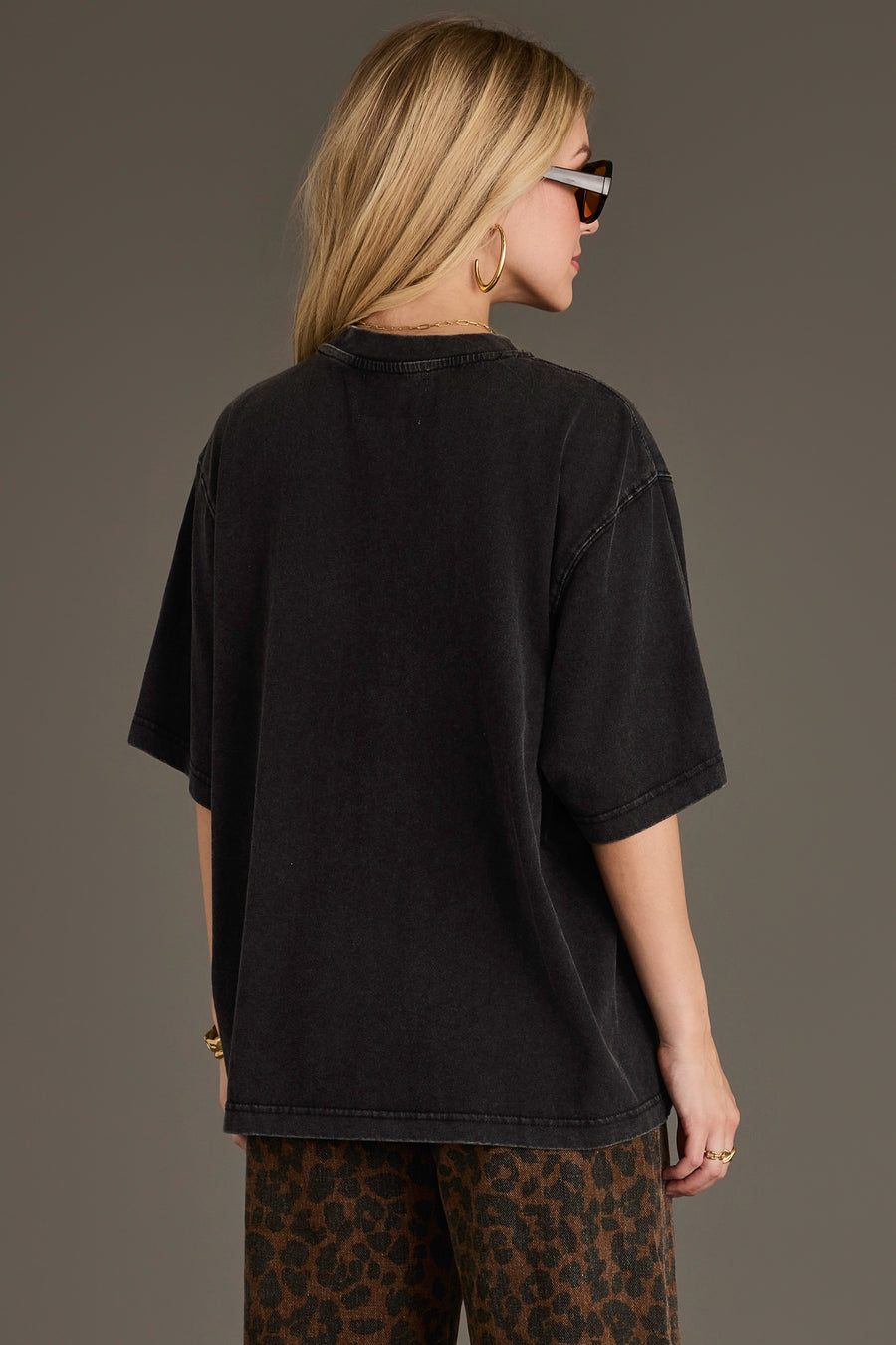 A person with long blonde hair, wearing sunglasses, gold hoop earrings, and leopard print pants, faces away from the camera against a plain background while dressed in the Panache Apparel Collection Black Whiskey Weather Tee, a relaxed fit washed black cotton top.