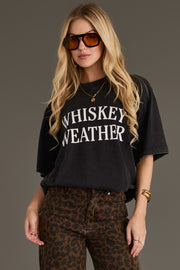 A woman with long blonde hair wears large brown sunglasses, leopard print pants, and gold jewelry, standing against a plain background. She is wearing the Panache Apparel Collection Black Whiskey Weather Tee in a relaxed fit.