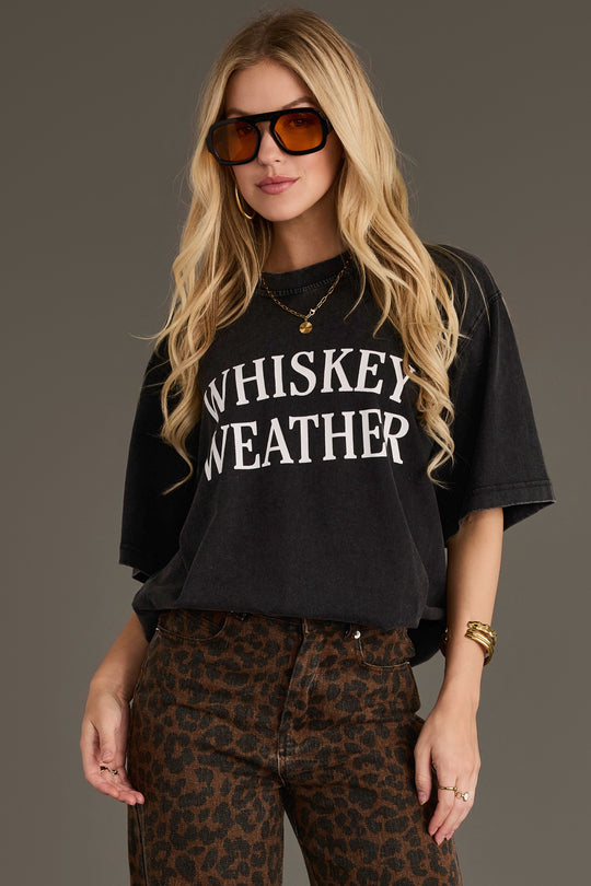 A woman with long blonde hair wears large brown sunglasses, leopard print pants, and gold jewelry, standing against a plain background. She is wearing the Panache Apparel Collection Black Whiskey Weather Tee in a relaxed fit.
