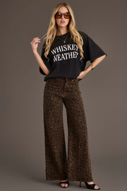 A woman wearing sunglasses and gold jewelry stands confidently against a plain background, styled in the Panache Apparel Collection Black Whiskey Weather Tee, a relaxed fit washed black cotton shirt featuring a bold “WHISKEY WEATHER?” print, paired with wide-leg leopard print pants and black heels.