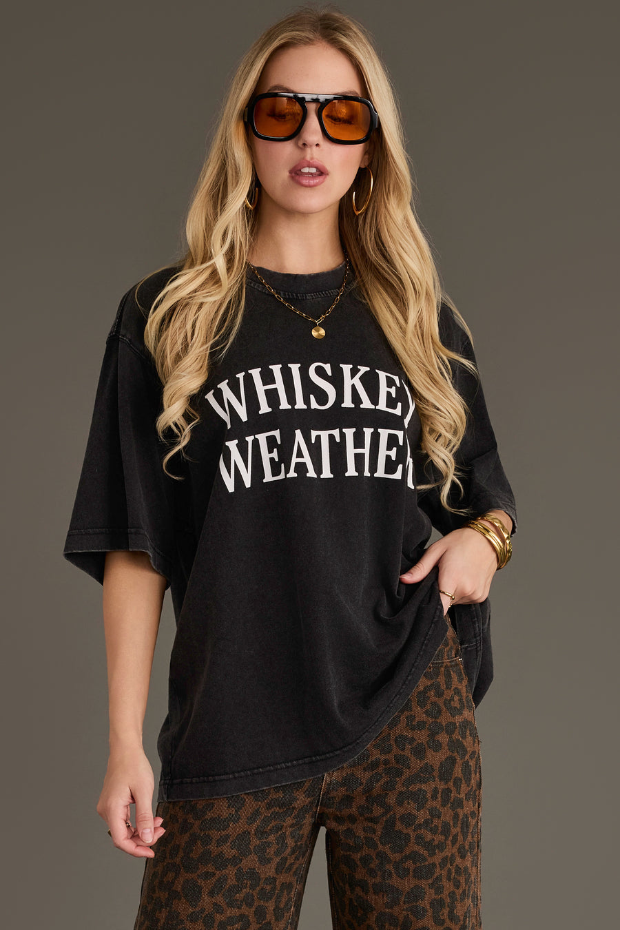 A woman with long blonde hair wears sunglasses, hoop earrings, the Panache Apparel Collection Black Whiskey Weather Tee, and leopard print pants, posing against a plain background.