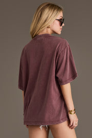 A woman with long blonde hair wears sunglasses and the Wine Western List Tee from Panache Apparel Collection in an oversized fit, paired with frayed denim shorts. She is facing away from the camera against a plain background.