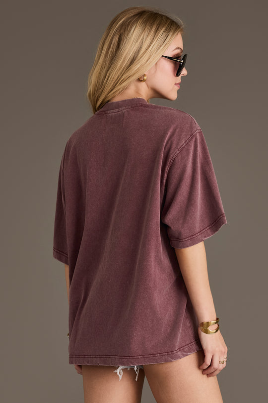 A woman with long blonde hair wears sunglasses and the Wine Western List Tee from Panache Apparel Collection in an oversized fit, paired with frayed denim shorts. She is facing away from the camera against a plain background.