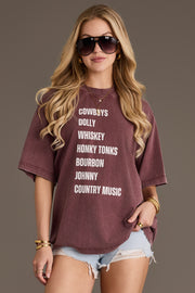 A woman with long blonde hair wears sunglasses, denim shorts, and the Panache Apparel Collection’s Wine Western List Tee. She stands against a plain background, holding the bottom of her shirt with one hand. The breathable cotton fabric and oversized fit of this tee give her outfit a relaxed and comfortable vibe.