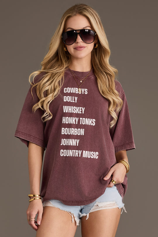 A woman with long blonde hair wears sunglasses, denim shorts, and the Panache Apparel Collection’s Wine Western List Tee. She stands against a plain background, holding the bottom of her shirt with one hand. The breathable cotton fabric and oversized fit of this tee give her outfit a relaxed and comfortable vibe.