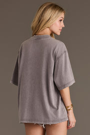 A woman with blonde hair wears the Panache Apparel Collection Cowboy Country Tee—an oversized, short-sleeve gray cotton t-shirt—paired with light denim shorts. She stands sideways and faces away, her head turned slightly to the right.