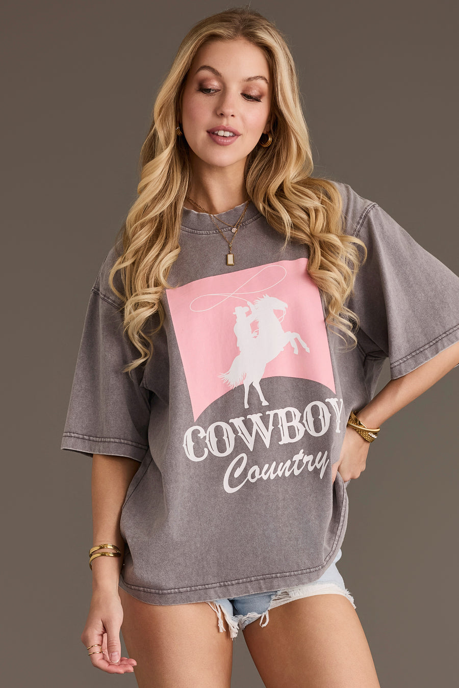 A woman with long blonde hair is wearing the Panache Apparel Collection Cowboy Country Tee—a gray cotton oversized graphic top featuring a pink and white lasso design—paired with denim shorts and gold jewelry, standing against a plain background.
