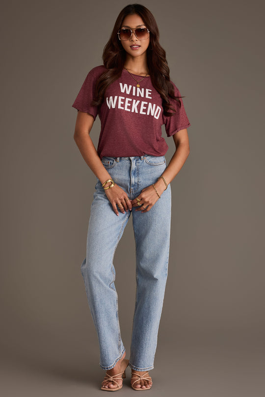 A person wearing a burgundy Wine Weekend Tee paired with light-wash jeans and heeled sandals.