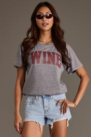 A person wearing a Panache Apparel Collection Gray Wine Tee featuring the word WINE in large maroon letters across the chest, paired with light-wash distressed denim shorts.