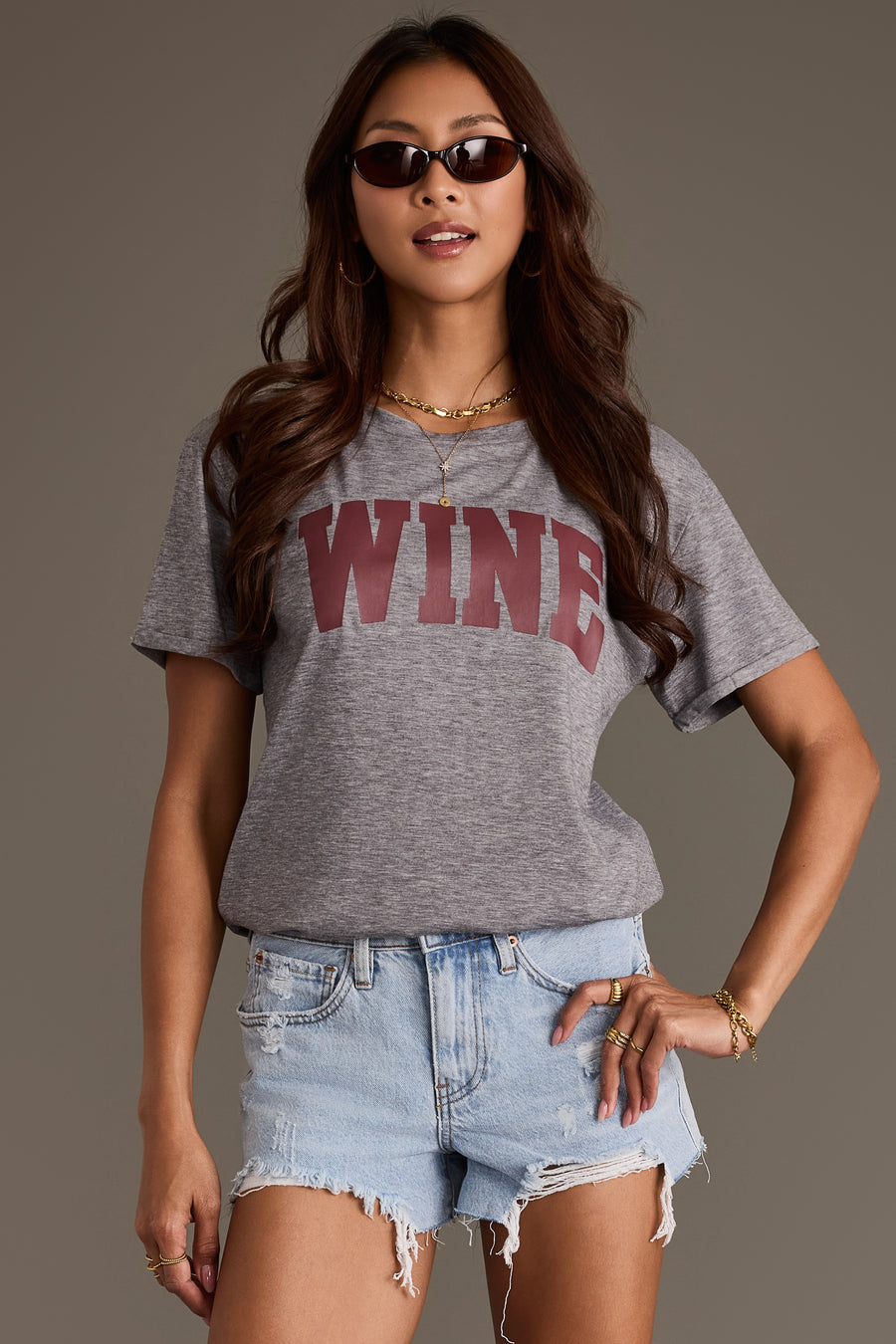 A person wearing a Panache Apparel Collection Gray Wine Tee featuring the word WINE in large maroon letters across the chest, paired with light-wash distressed denim shorts.