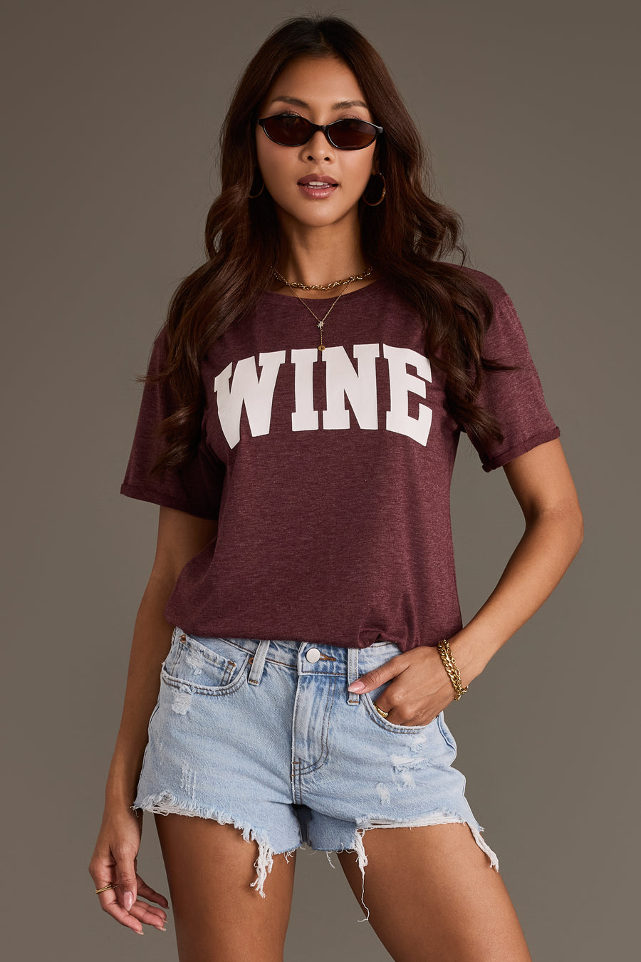 A person wearing a Burgundy Wine Tee with the word WINE printed in white block letters, paired with distressed light-wash denim shorts.