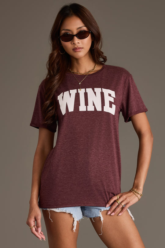 A person wearing sunglasses and a relaxed fit Burgundy Wine Tee from Panache Apparel Collection with large white letters, paired with denim shorts.