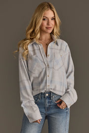 A woman with long blonde hair, dressed in the Panache Apparel Collection’s White, Gray & Light Blue Plaid Button Up and blue jeans, stands against a plain gray background with a slight smile.