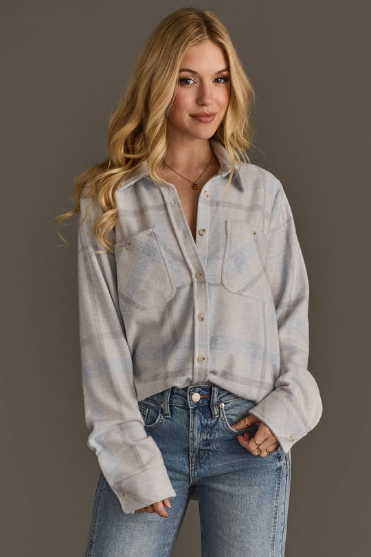 A woman with long blonde hair, dressed in the Panache Apparel Collection’s White, Gray & Light Blue Plaid Button Up and blue jeans, stands against a plain gray background with a slight smile.