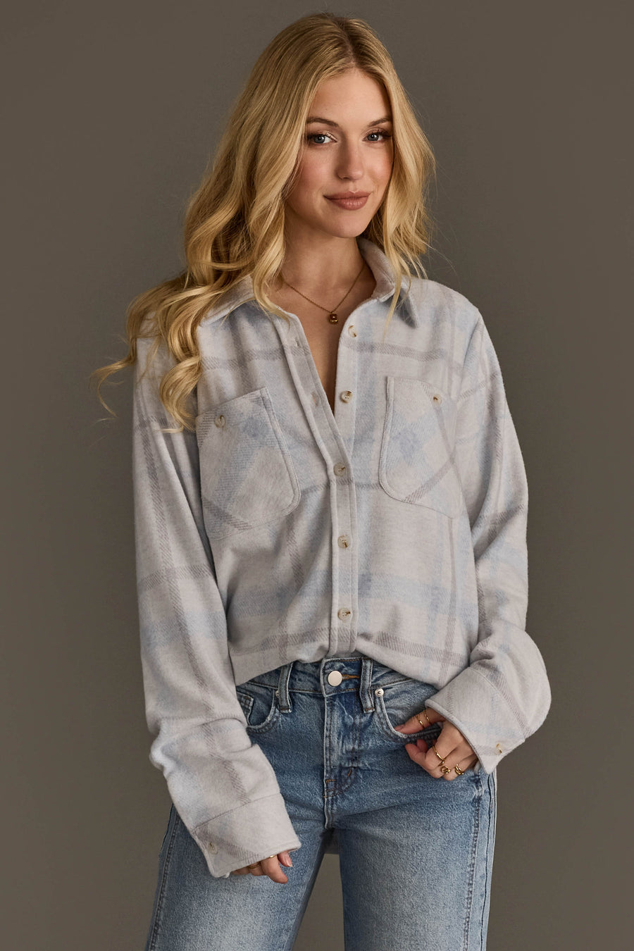 A woman with long blonde hair wears the White, Gray & Light Blue Plaid Button Up from Panache Apparel Collection and blue jeans, standing against a plain gray background and smiling softly.
