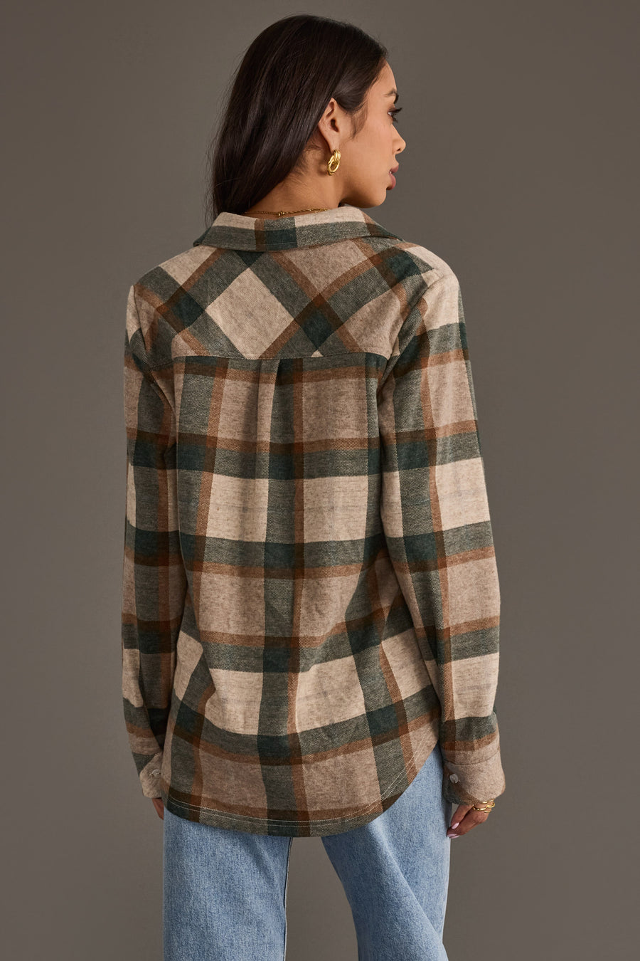 A woman stands facing away, wearing the Panache Apparel Collection Tan, Dark Green & Rust Plaid Button Up with blue jeans. She has long dark hair, gold hoop earrings, and the background is plain gray.
