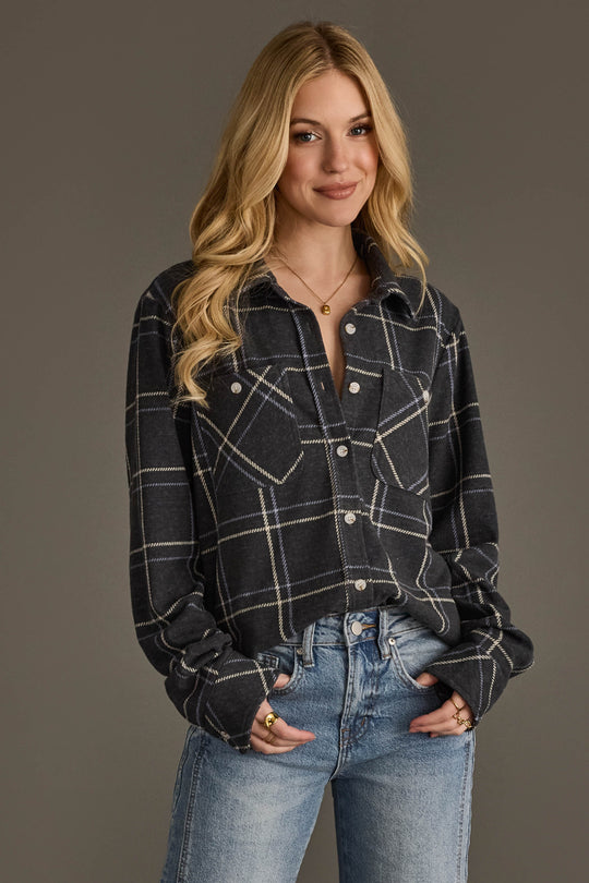 A woman with long blonde hair stands against a plain background, smiling with her hands in her pockets while wearing the Panache Apparel Collection Dark Gray, Light Blue & White Plaid Button Up and blue jeans.